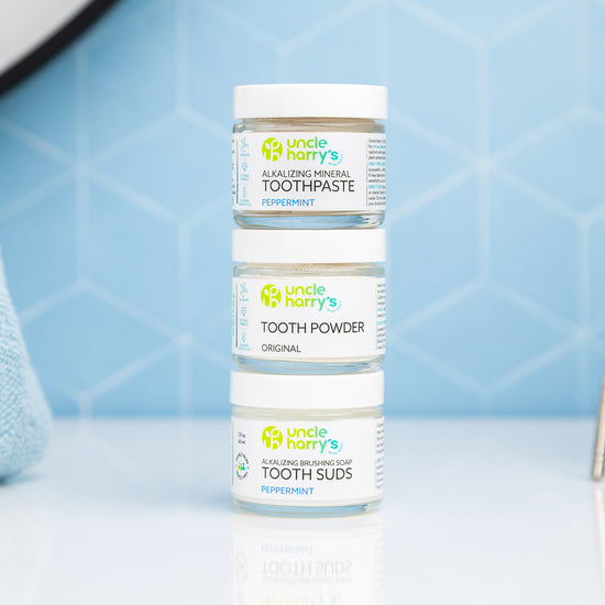 Bottles of Uncle Harry's Toothpaste, Tooth Powder, Tooth Suds on a bathroom counter with a toothbrush and glass in the background.