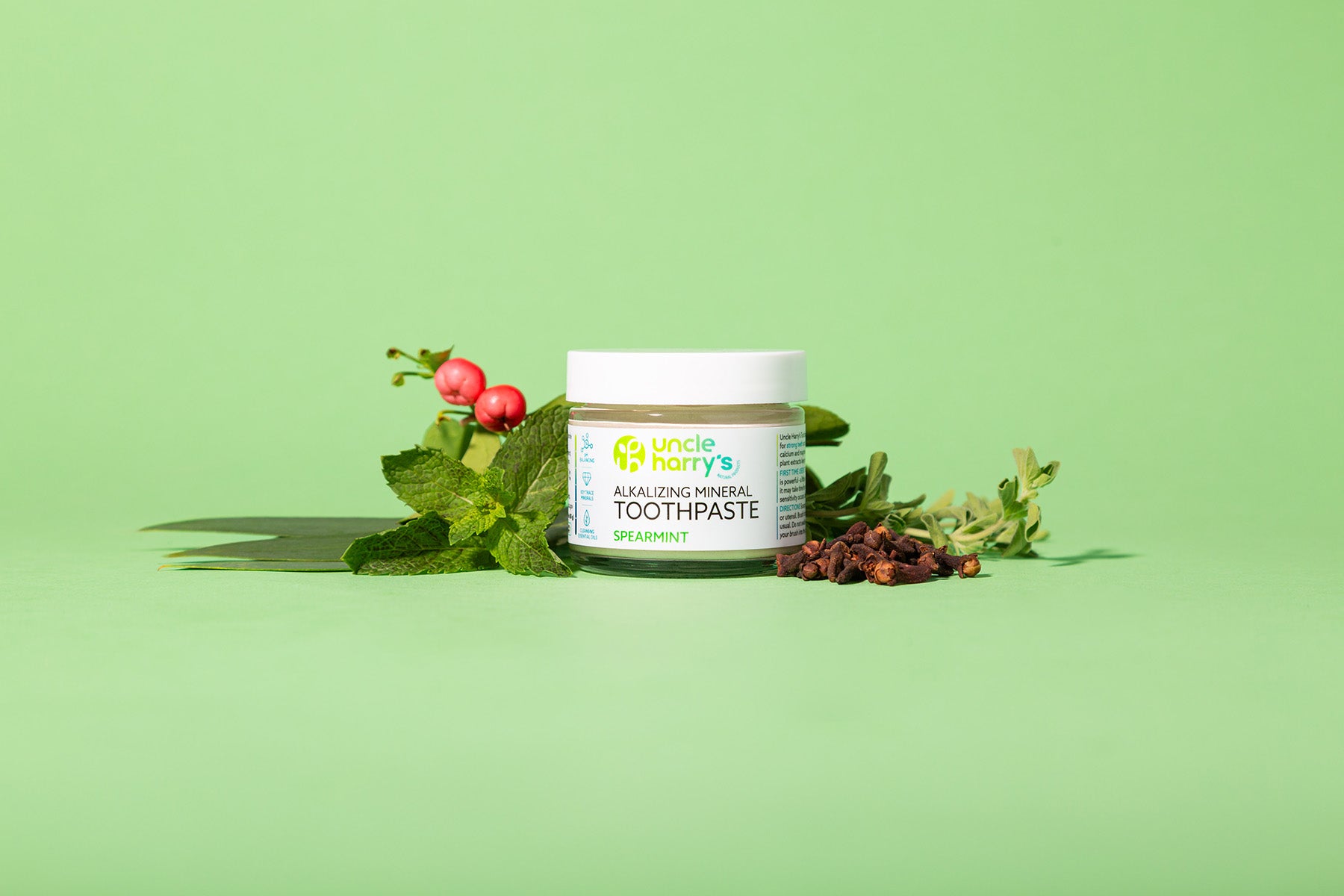 Jar of Uncle Harry's Natural Spearmint Toothpaste with mint leaves and berries on a green background